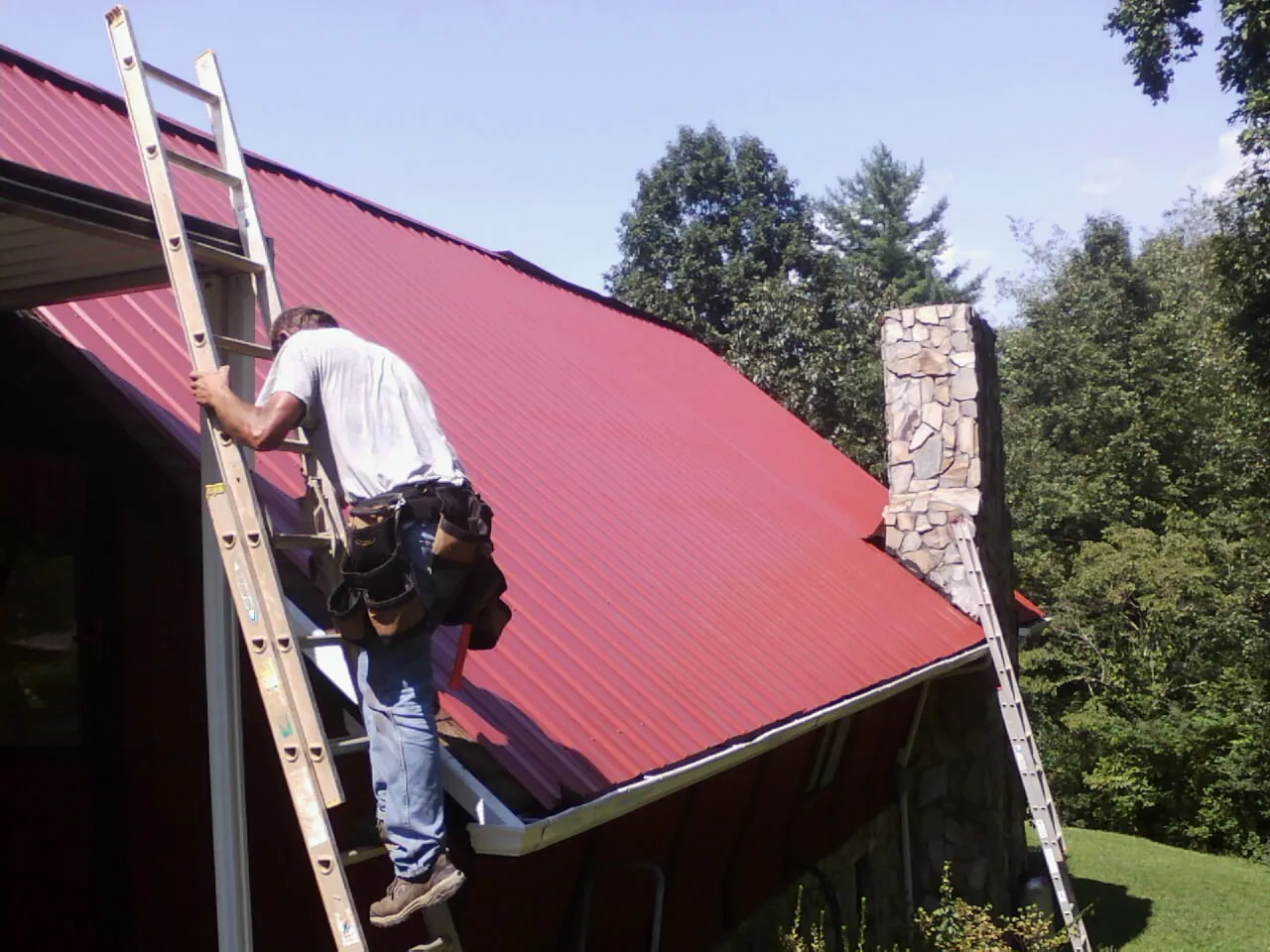 Roofer on a ladder beside a red metal roof and stone chimney — Soffit & Fascia Repair by Roofing Brothers in Ratliff City